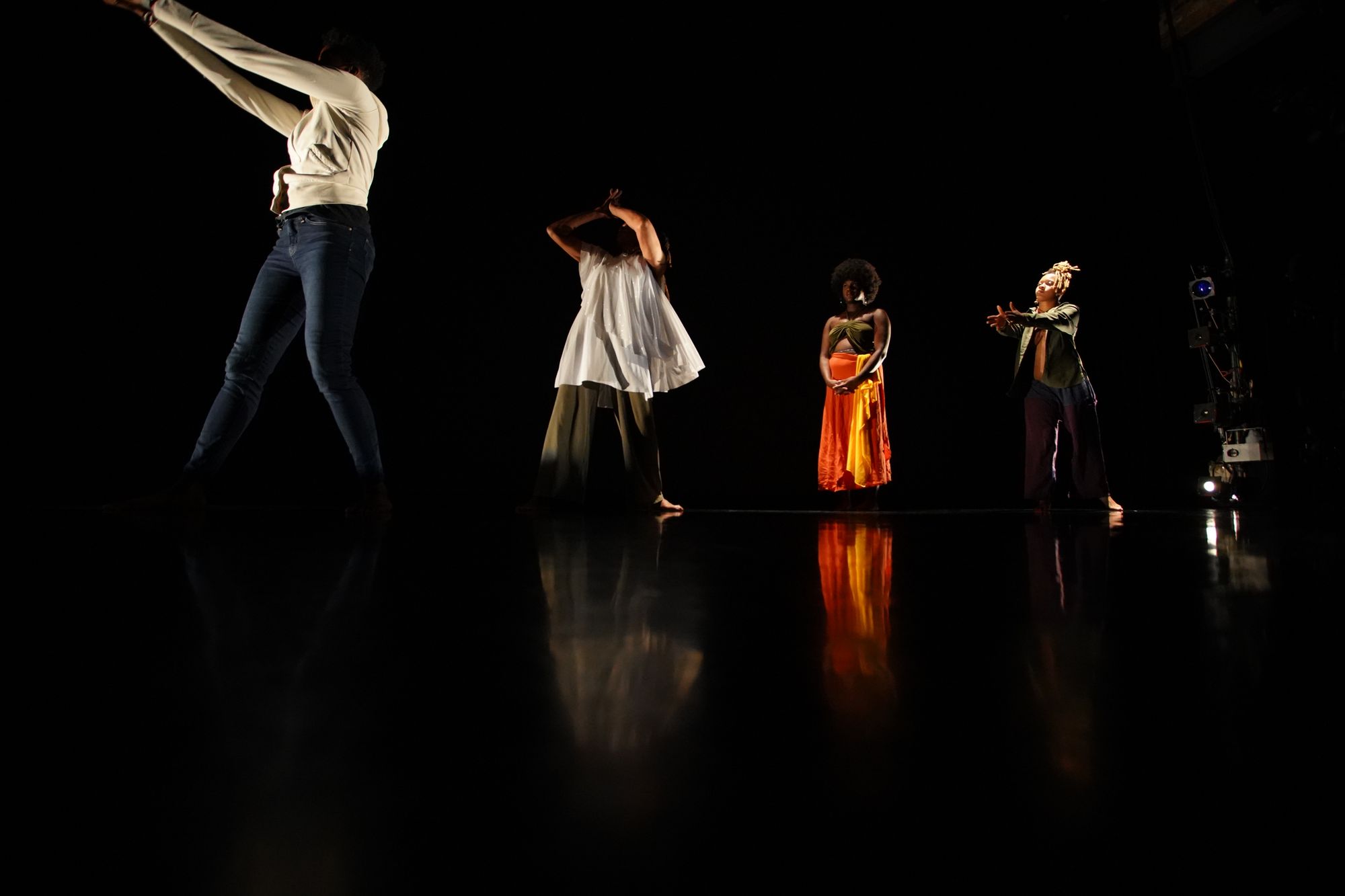 MK Abadoo, in blue jeans and a white jacket, has their right leg stepped forward with both arms extended up and in front. Enveloped in darkness, light beams from dancers Julinda Lewis, Christine Wyatt and Sehay Durant, as they emulate and witness MK’s movement.