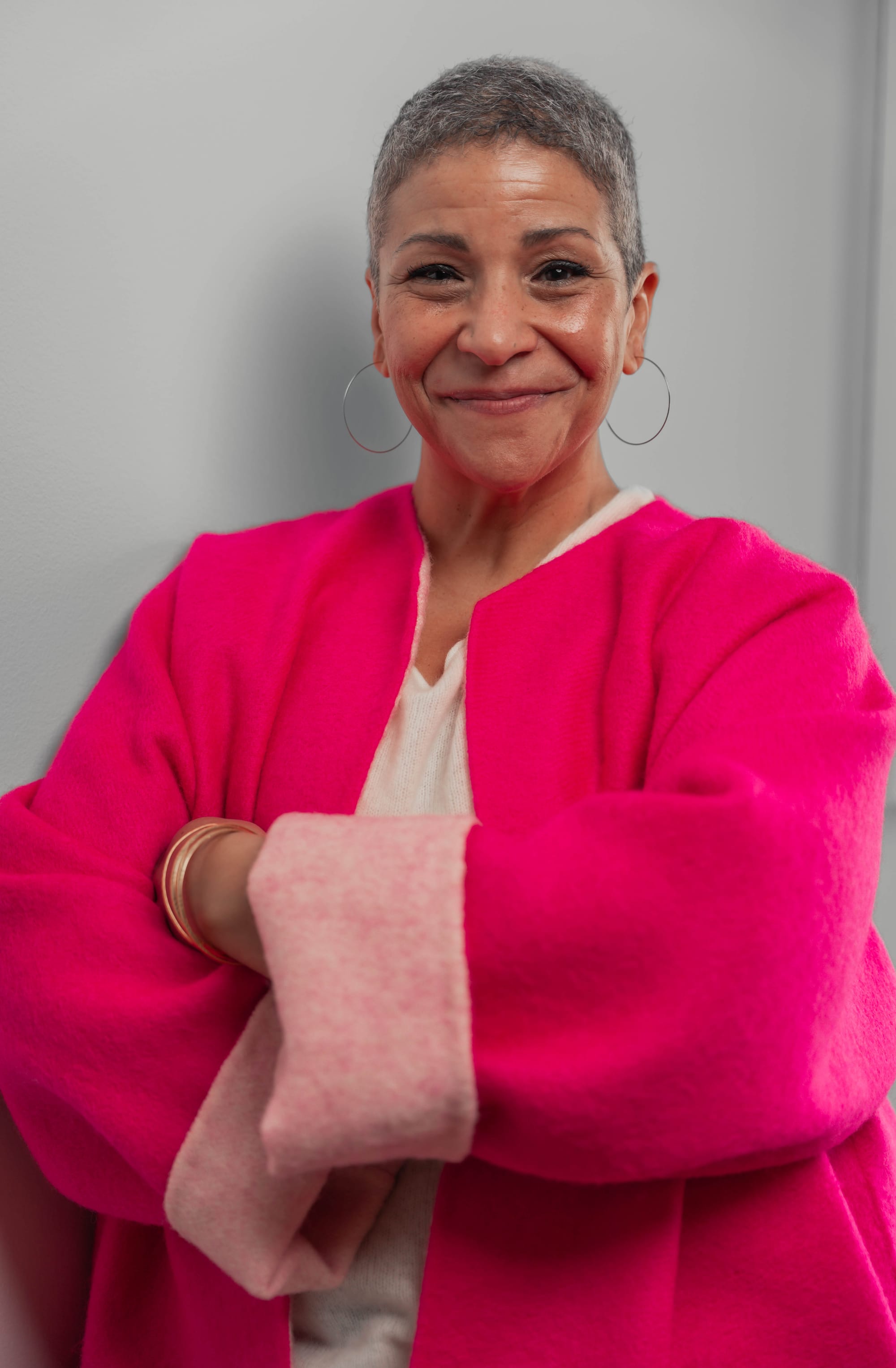 A smiling woman with short gray hair and silver hoop earrings wears a bright pink cardigan