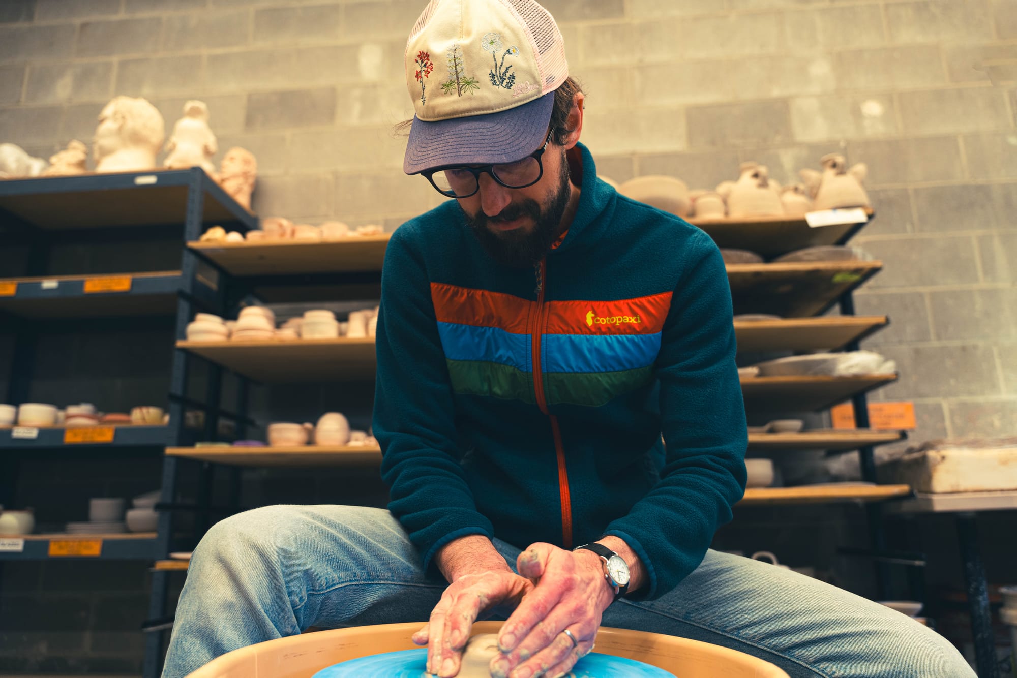 A man with glasses and floral baseball cap sits at a pottery wheel and molds clay