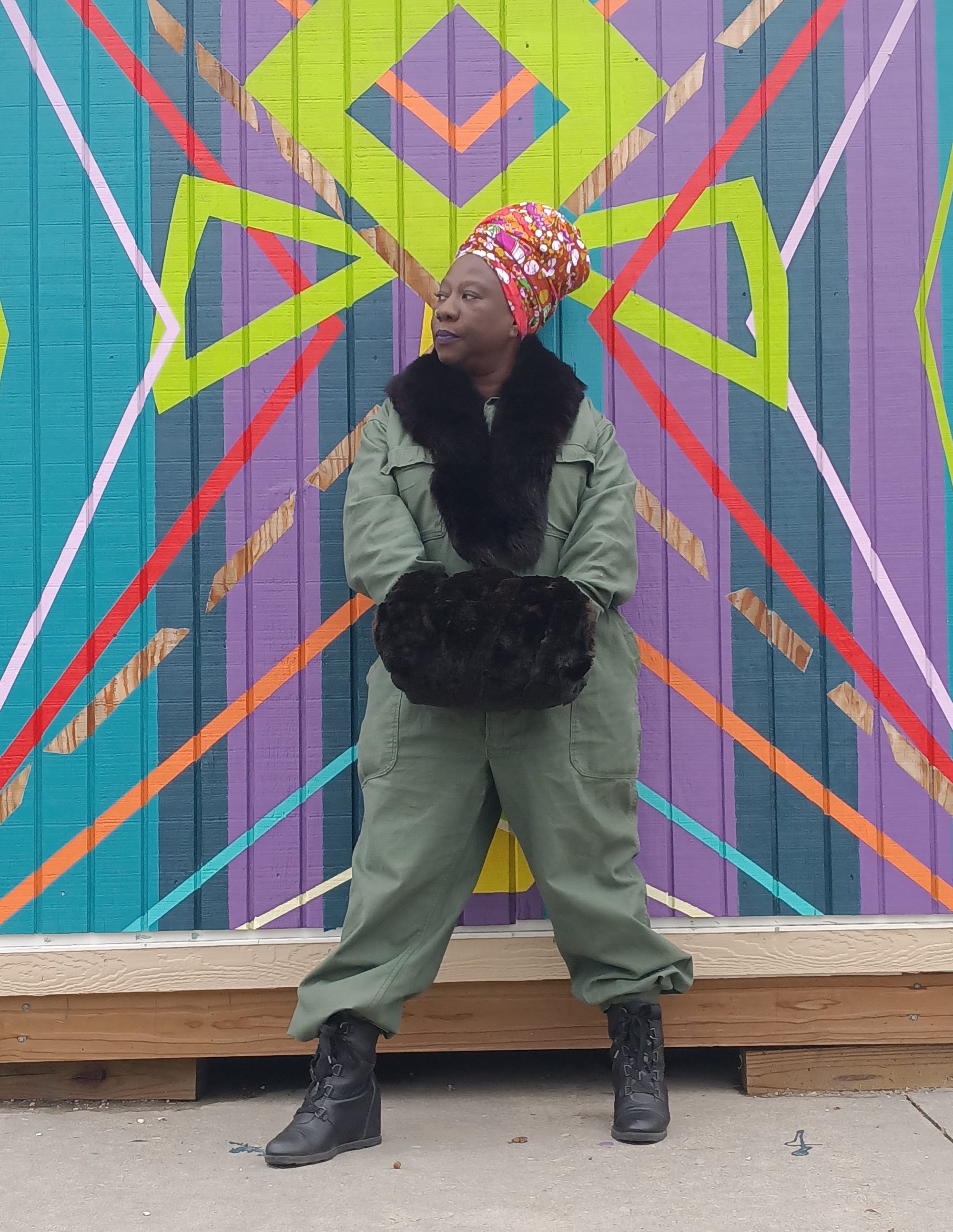 Celeste Butler wears a green jumpsuit and floral headwrap. She stands in front of a colorful wall outside.
