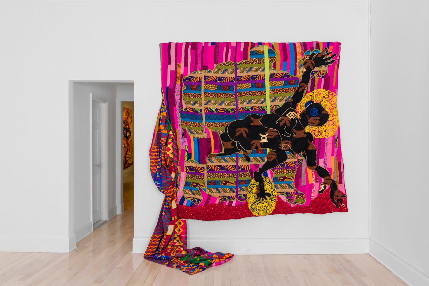 A-large-scale-quilt-with-a-black-figure-stretching-its-hands-outward-against-a-mainly-pink-and-red-and-purple-background.