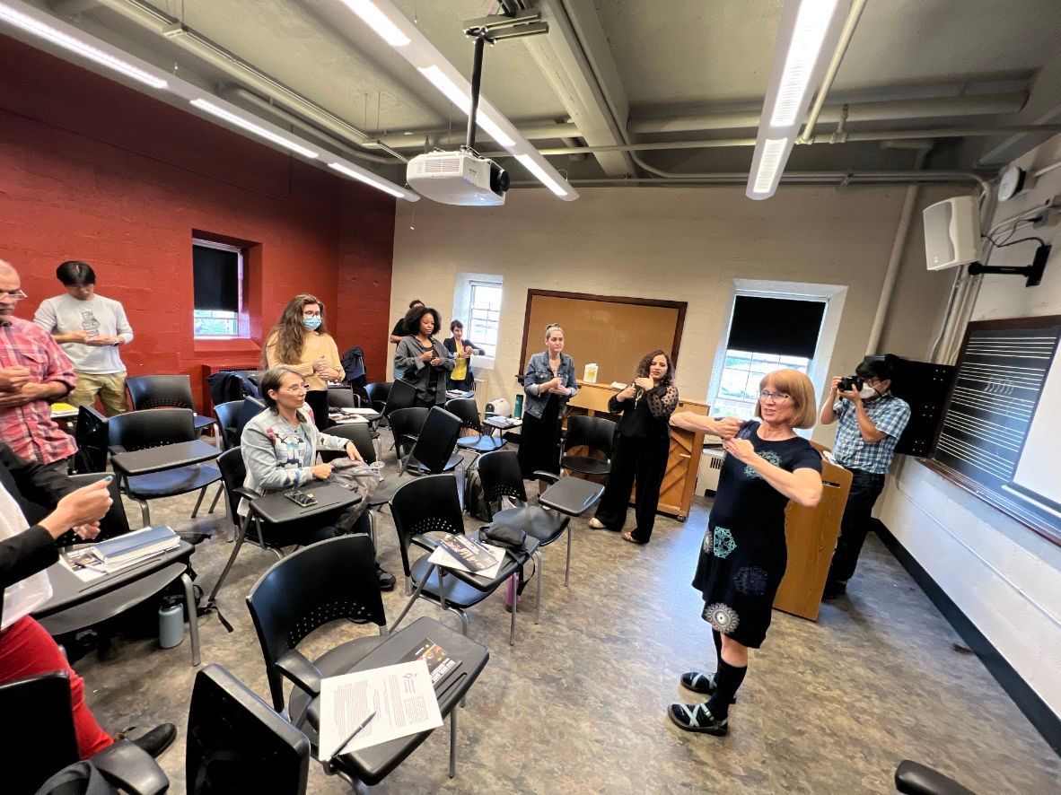 In-a-classroom-a-woman-demonstrates-how-to-play-carillon-using-her-fists.-There-are-9-others-in-the-classroom.