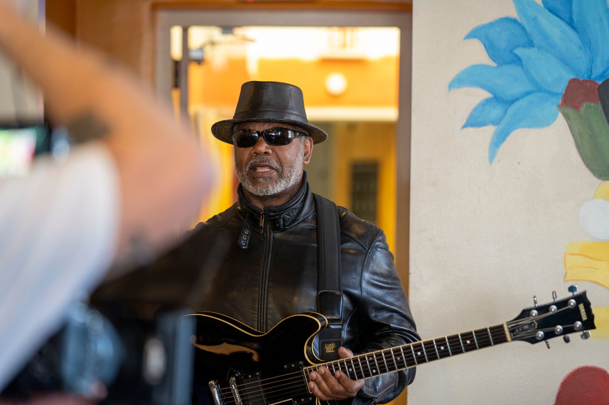 Image of a guitar player in a black leather jacket, sunglasses, and hat