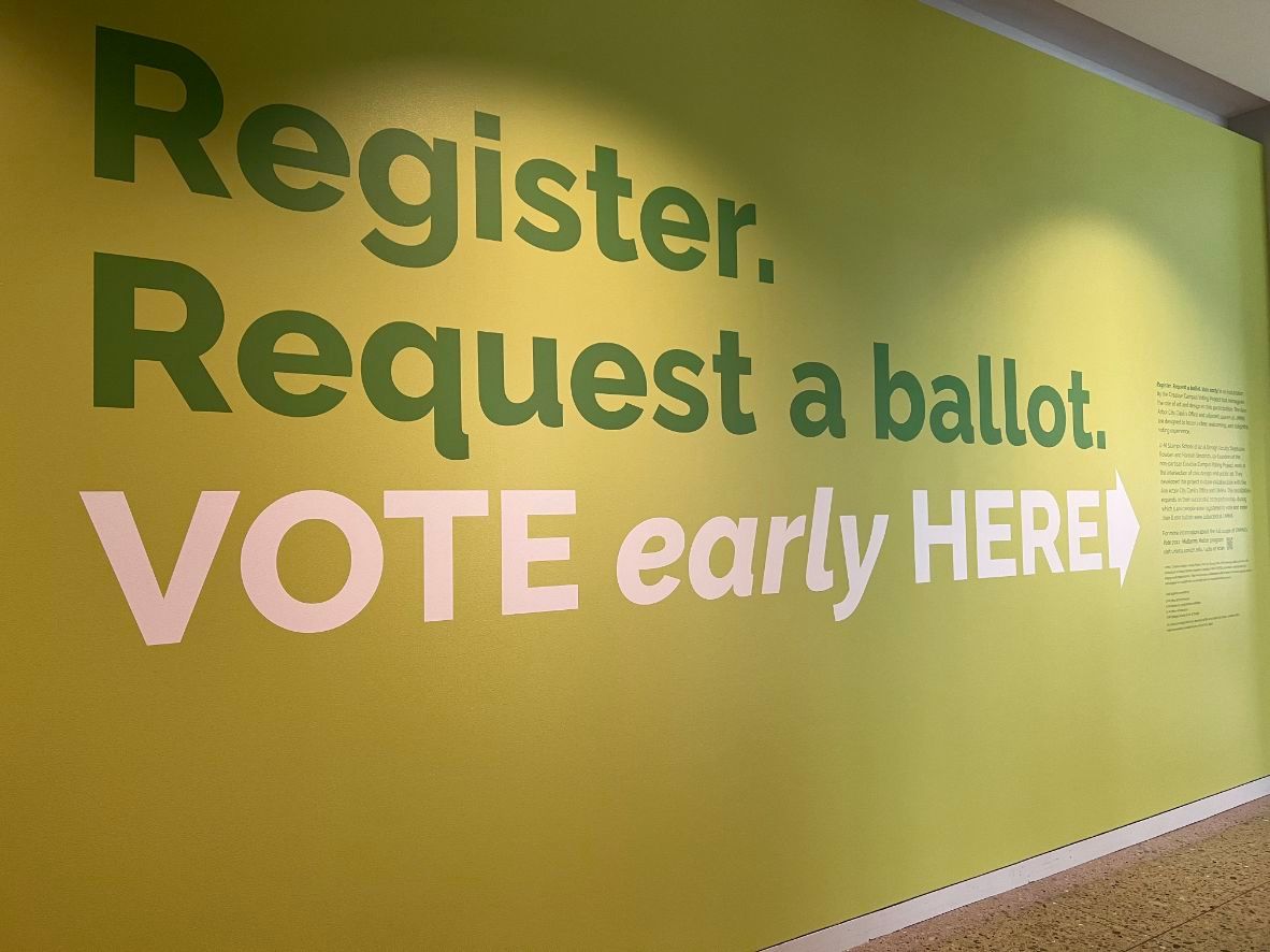 Large-text-on-a-gallery-wall-reads-register.-request-a-ballot.-vote-early-and-here.