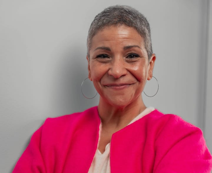 A woman with short gray hair and silver hoop earrings smiles. She's wearing a pink sweater