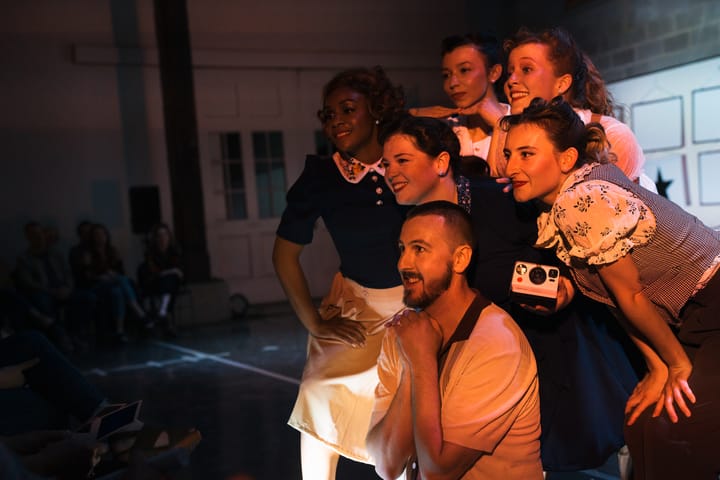 A group of six dancers dressed in 1950s attire dramatically pose for a photo on stage.